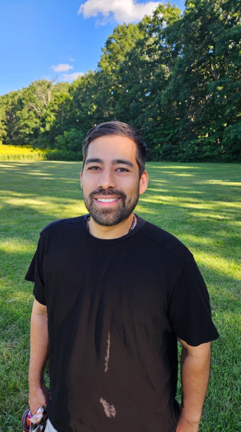 Nolan Fernandez smiling outdoors in a grassy meadow with trees behind him.